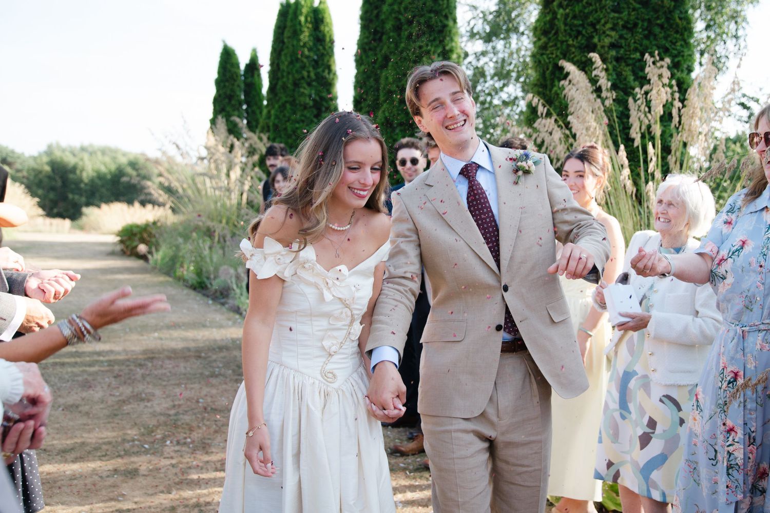 Bride and groom walk hand in hand through cheering wedding guests outdoors at a sunny rustic venue.