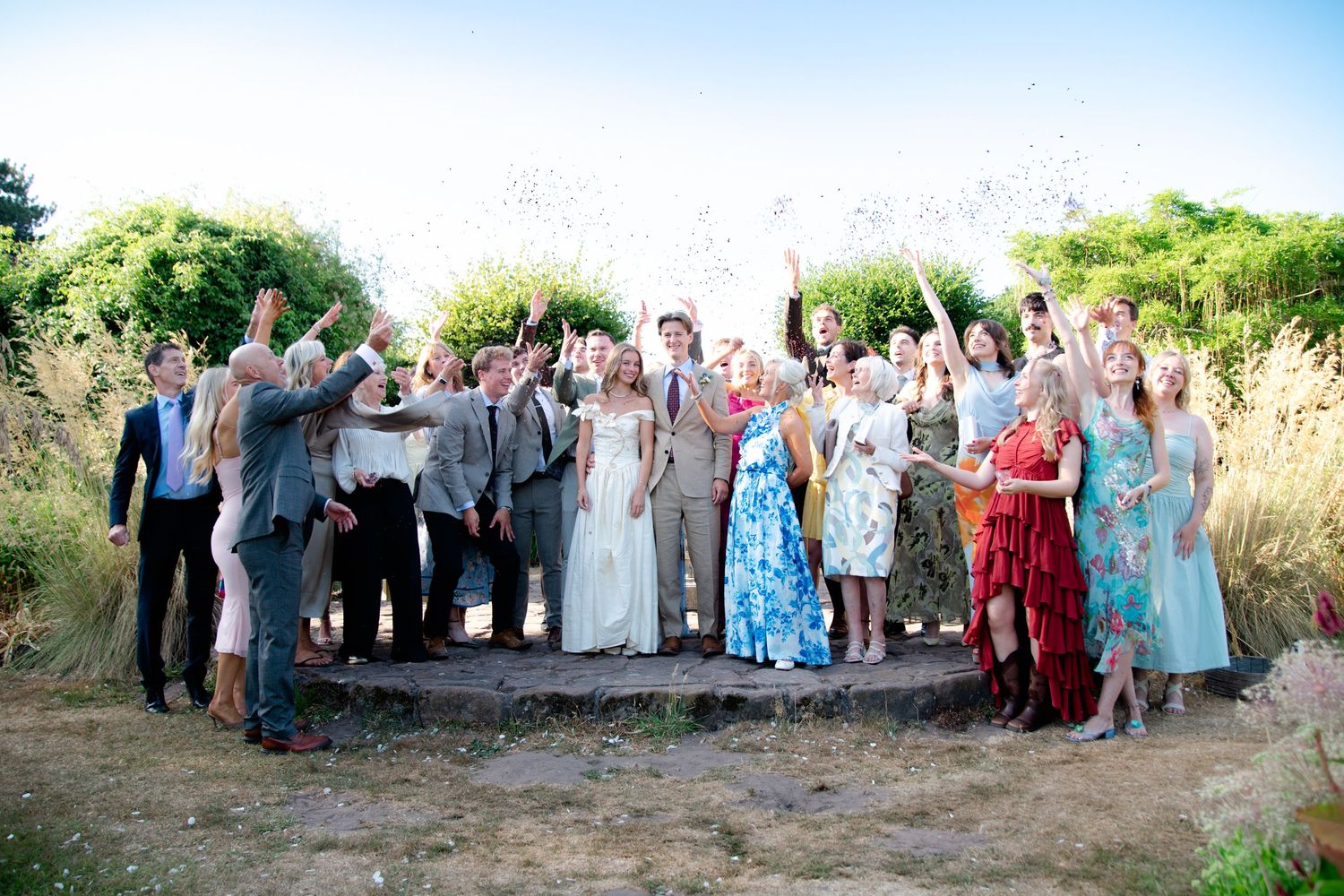 Wedding party celebrating outdoors, throwing confetti, bride in white gown surrounded by guests in colorful attire on a sunny day.