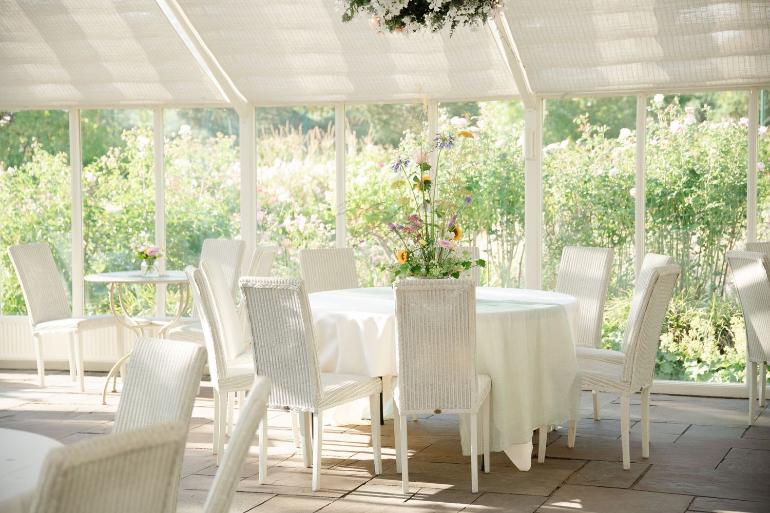 Bright sunlit conservatory dining room with white wicker chairs and round tables adorned with fresh floral centerpieces.