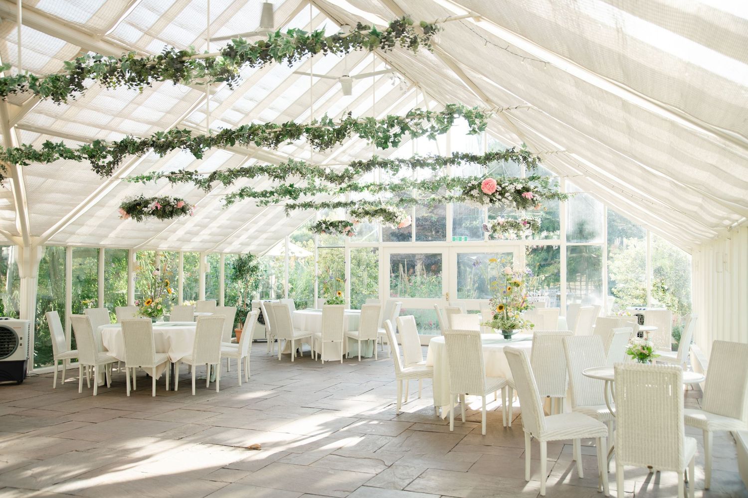 Elegant white conservatory wedding reception with floral garland ceiling, white chairs, and tables bathed in natural light.