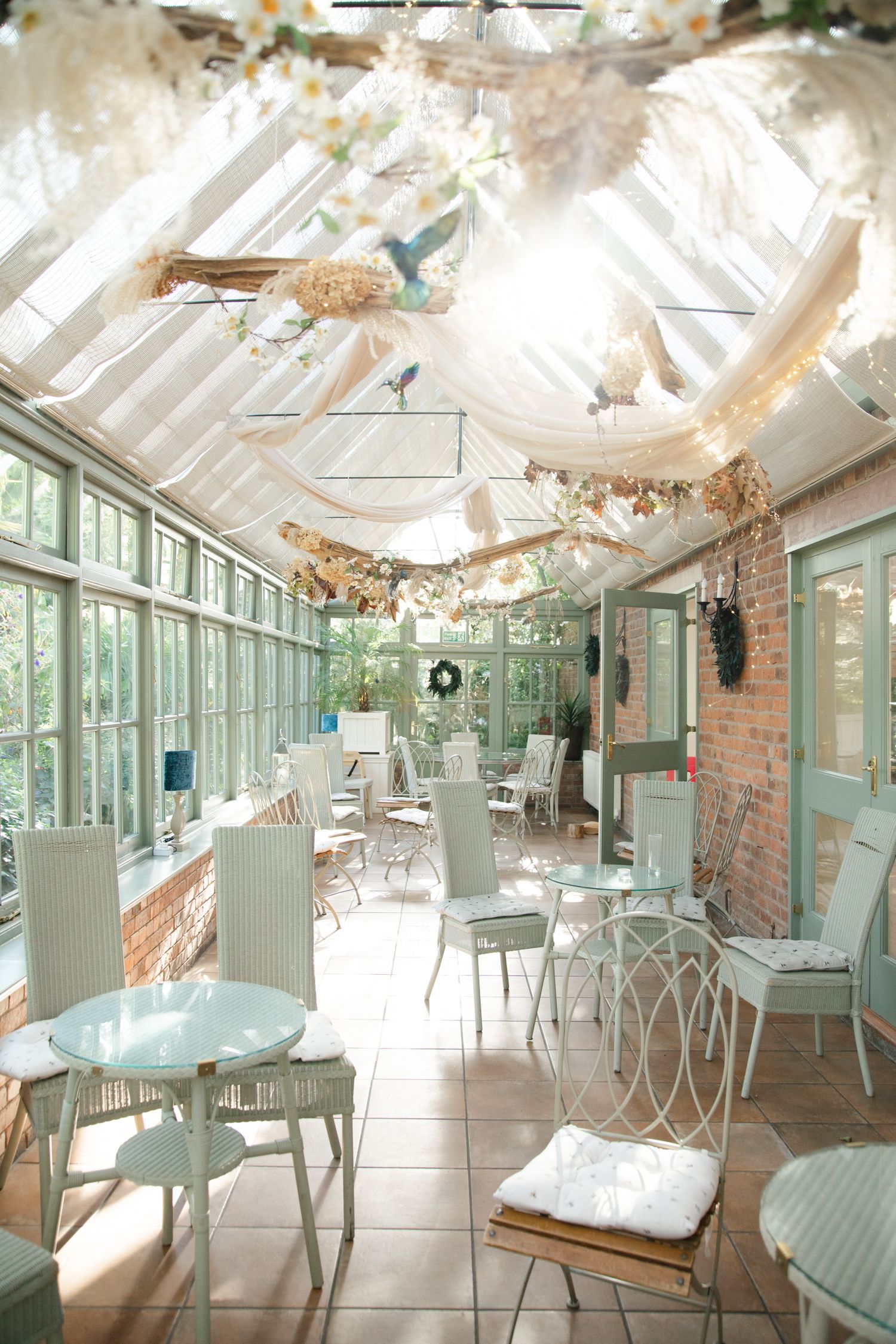 Bright sunroom cafe with white wicker chairs, glass ceiling, floral decor, and draped fabric in a charming garden setting.