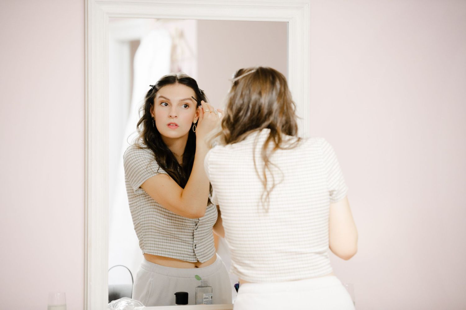 Young woman applying eye makeup while looking in a mirror, wearing a striped crop top in a bright room.