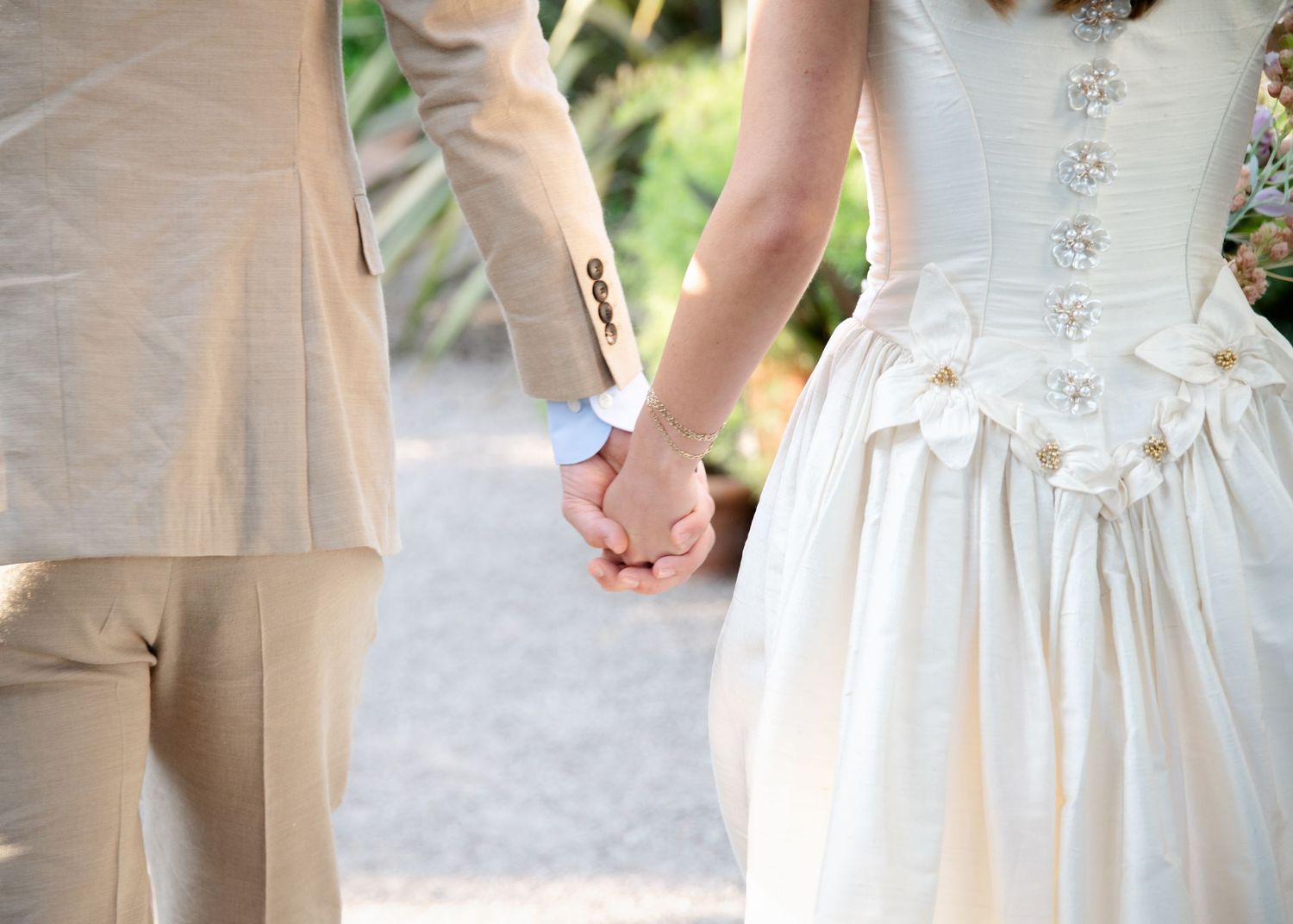 Bride and groom holding hands at outdoor wedding, woman in white corset dress, man in beige suit.