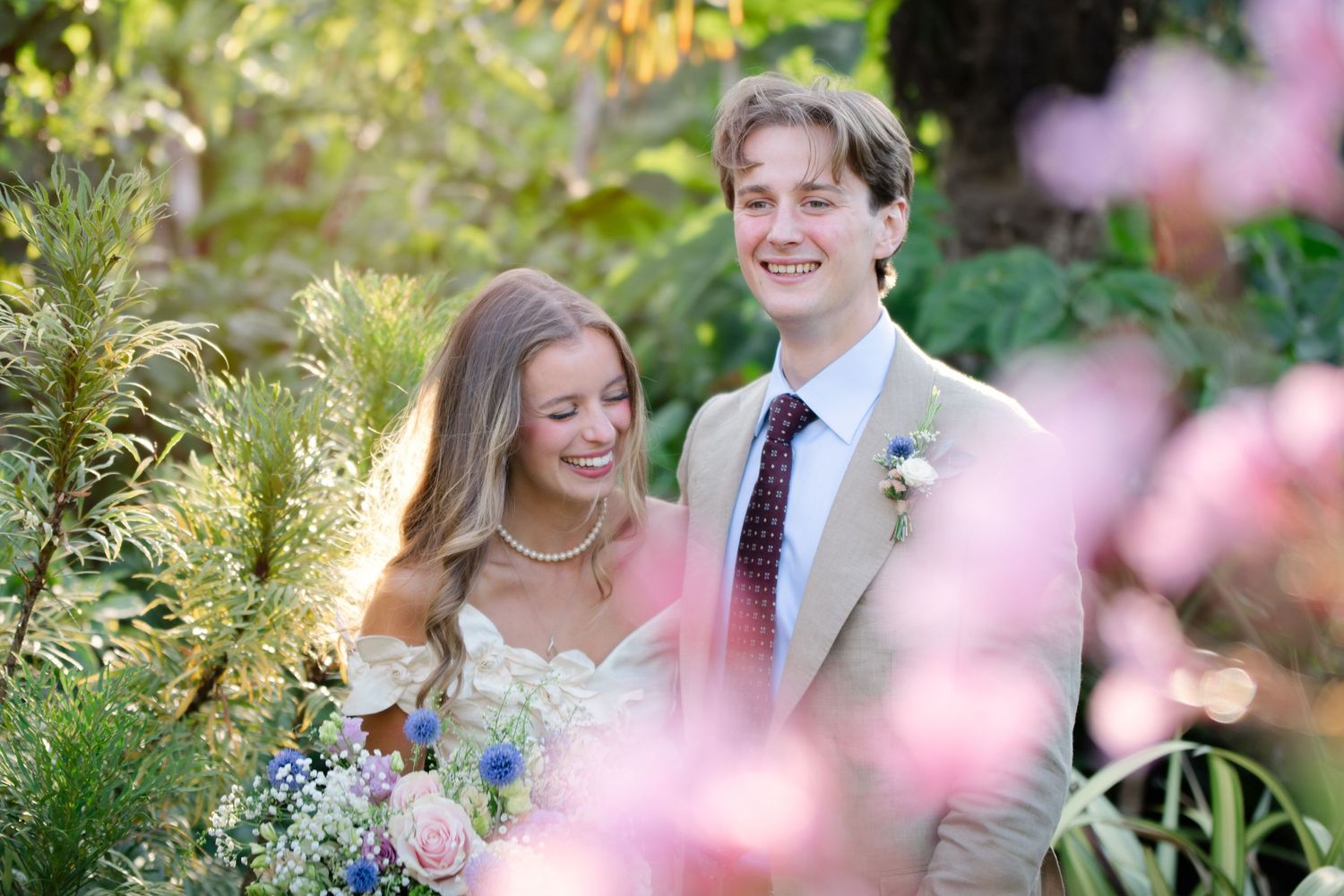 A smiling bride and groom pose among lush greenery and pink blossoms on their wedding day.