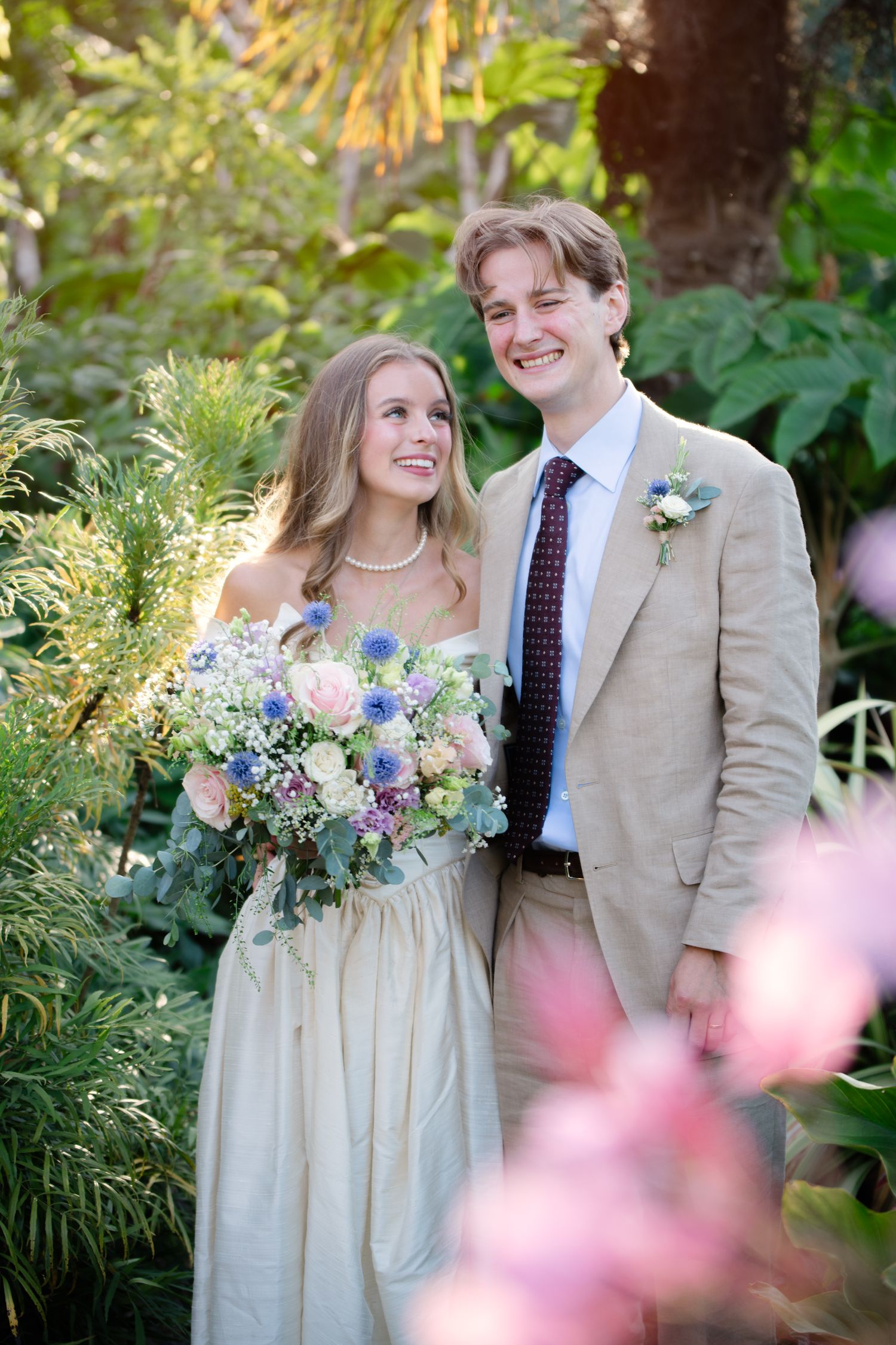 A smiling bride holding a pastel bouquet stands with a groom in a tan suit and boutonniere in a lush garden setting.