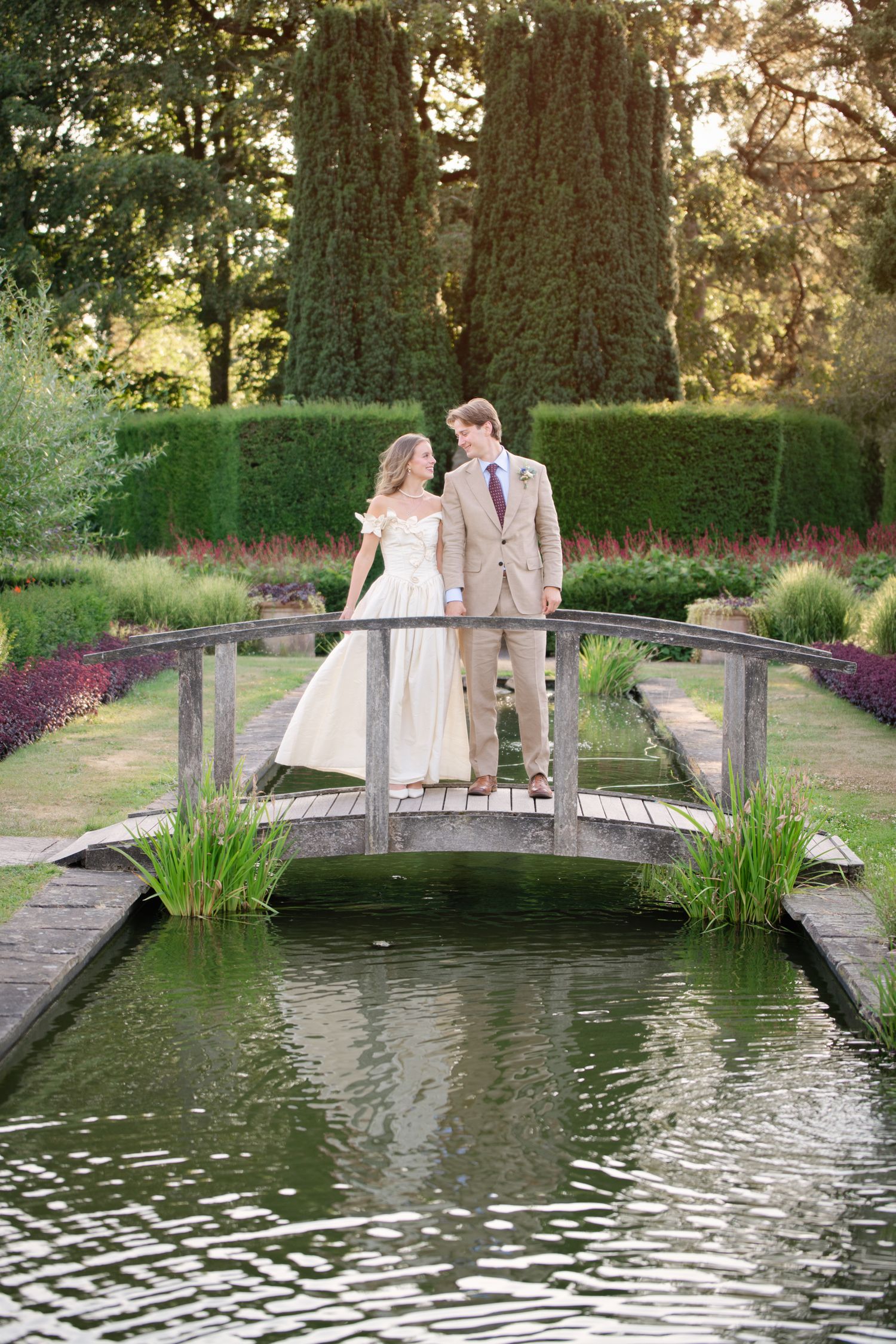Bride and groom stand on a wooden bridge over a pond in a lush garden, gazing at each other on their wedding day.