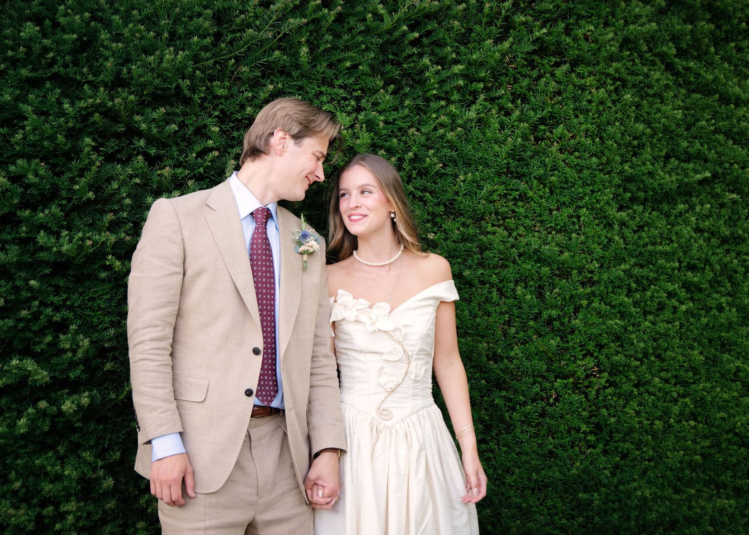 Couple in wedding attire smiling at each other in front of a lush green hedge wall.