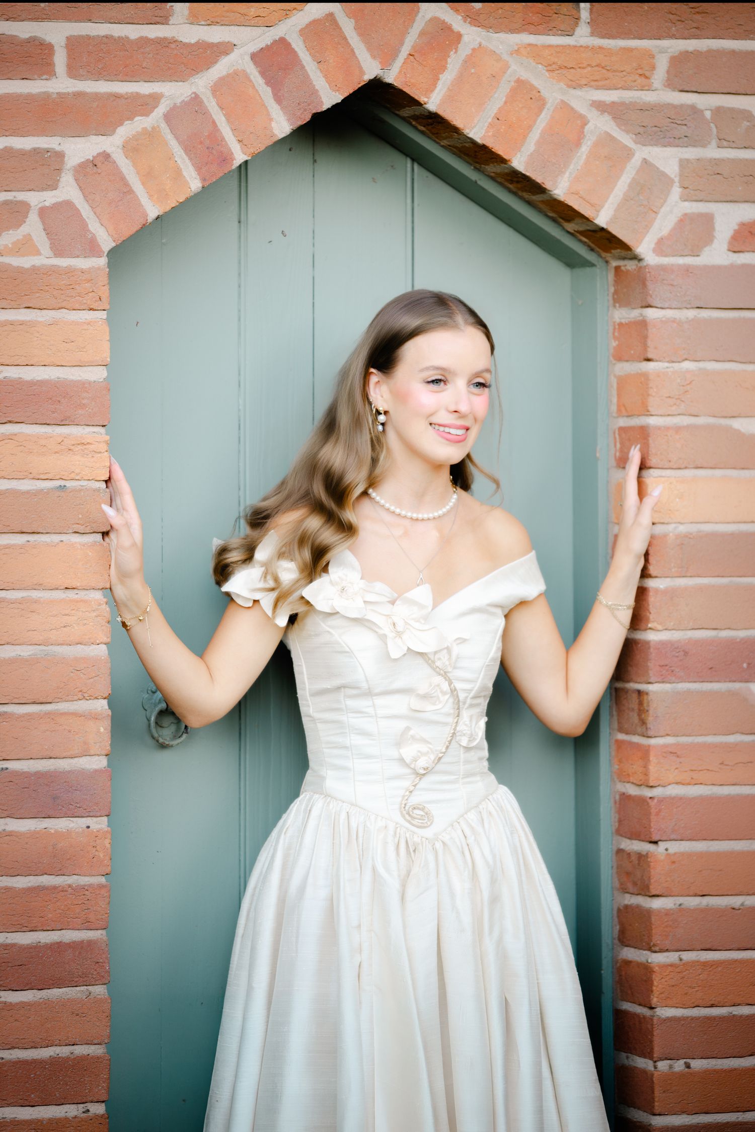 Young woman in elegant off-shoulder white gown smiling in a brick archway doorway.