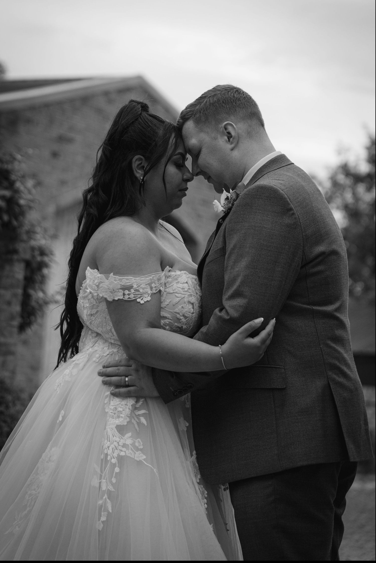 Black and white wedding photo of bride in off-shoulder gown and groom in suit sharing an intimate moment outdoors.