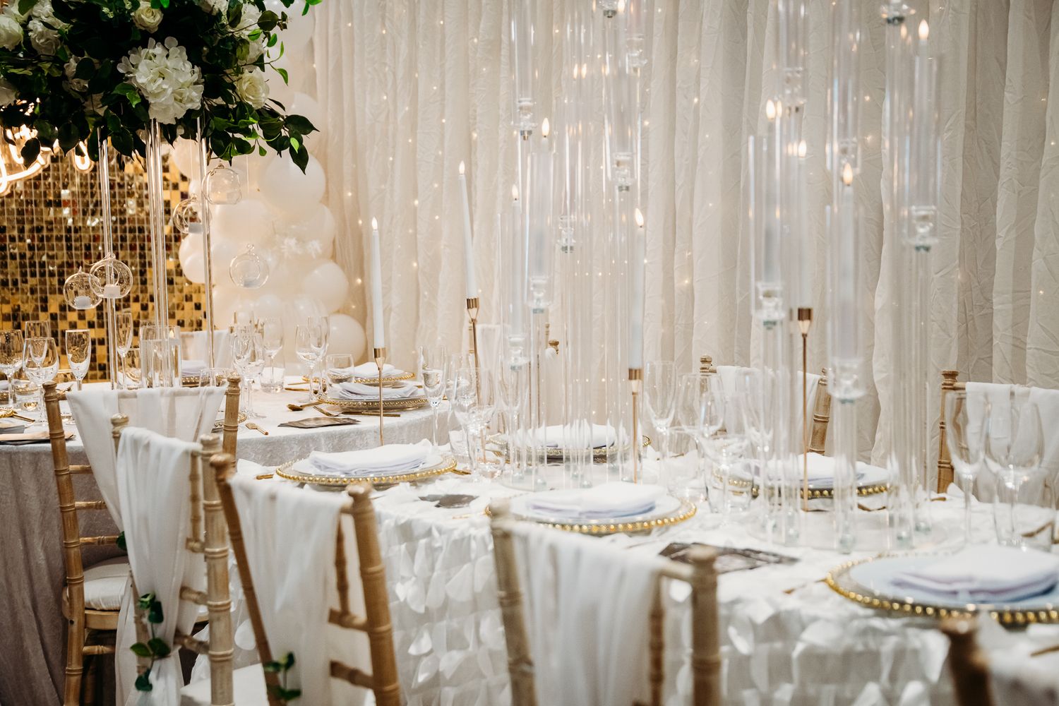 Elegant wedding reception table with white draping, gold chiavari chairs, crystal glassware, and floral centerpieces.