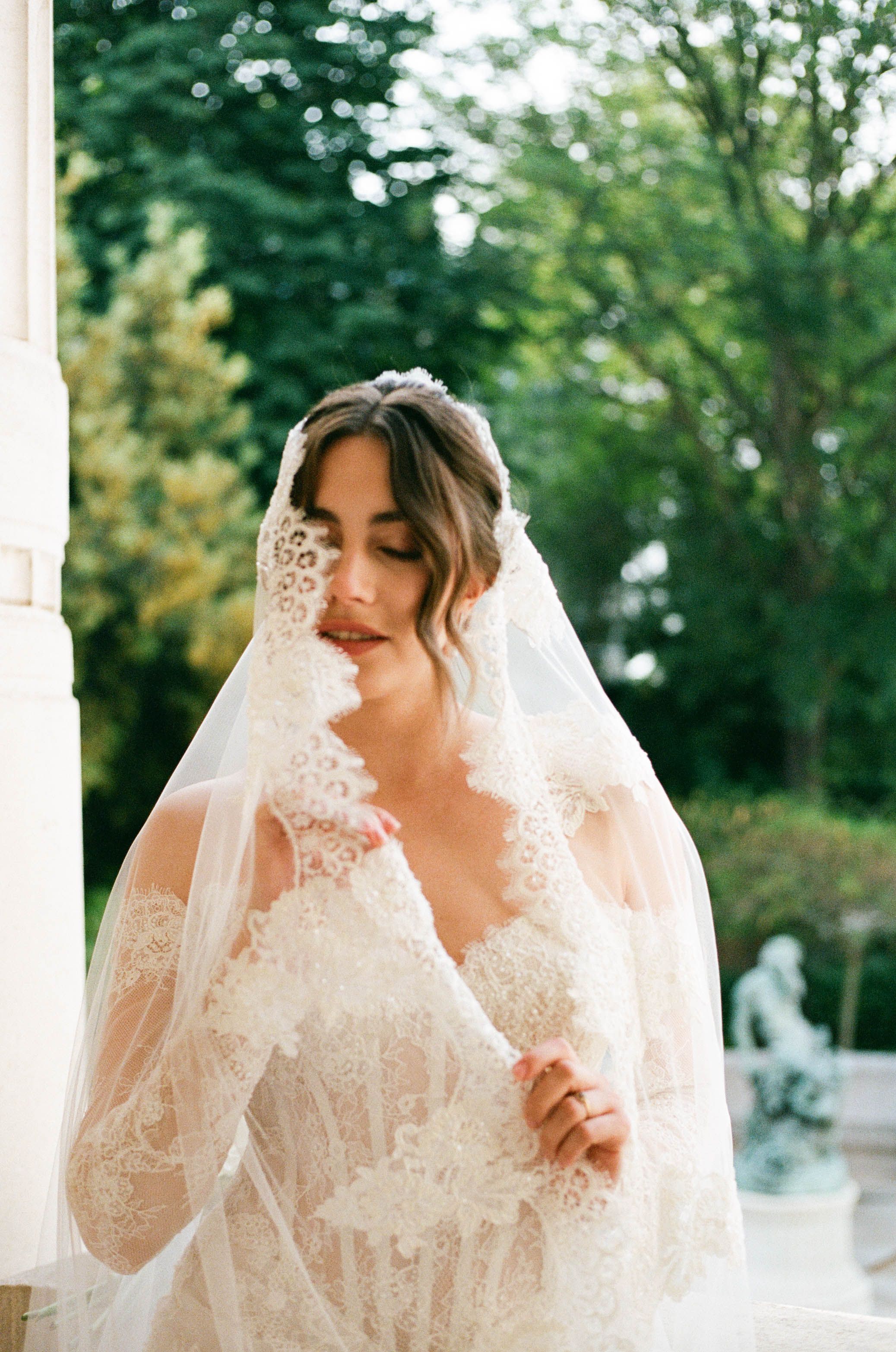 Bride in lace wedding dress and veil poses outdoors with lush green trees in soft natural light.