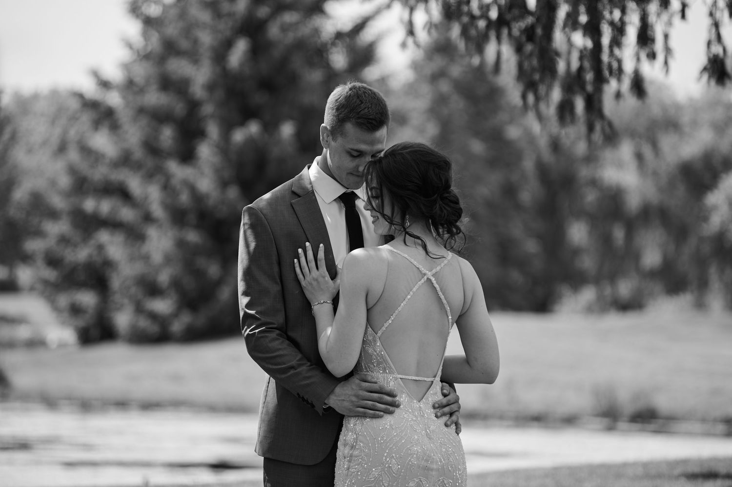 Black and white wedding photo of bride and groom sharing an intimate moment outdoors among lush green trees.