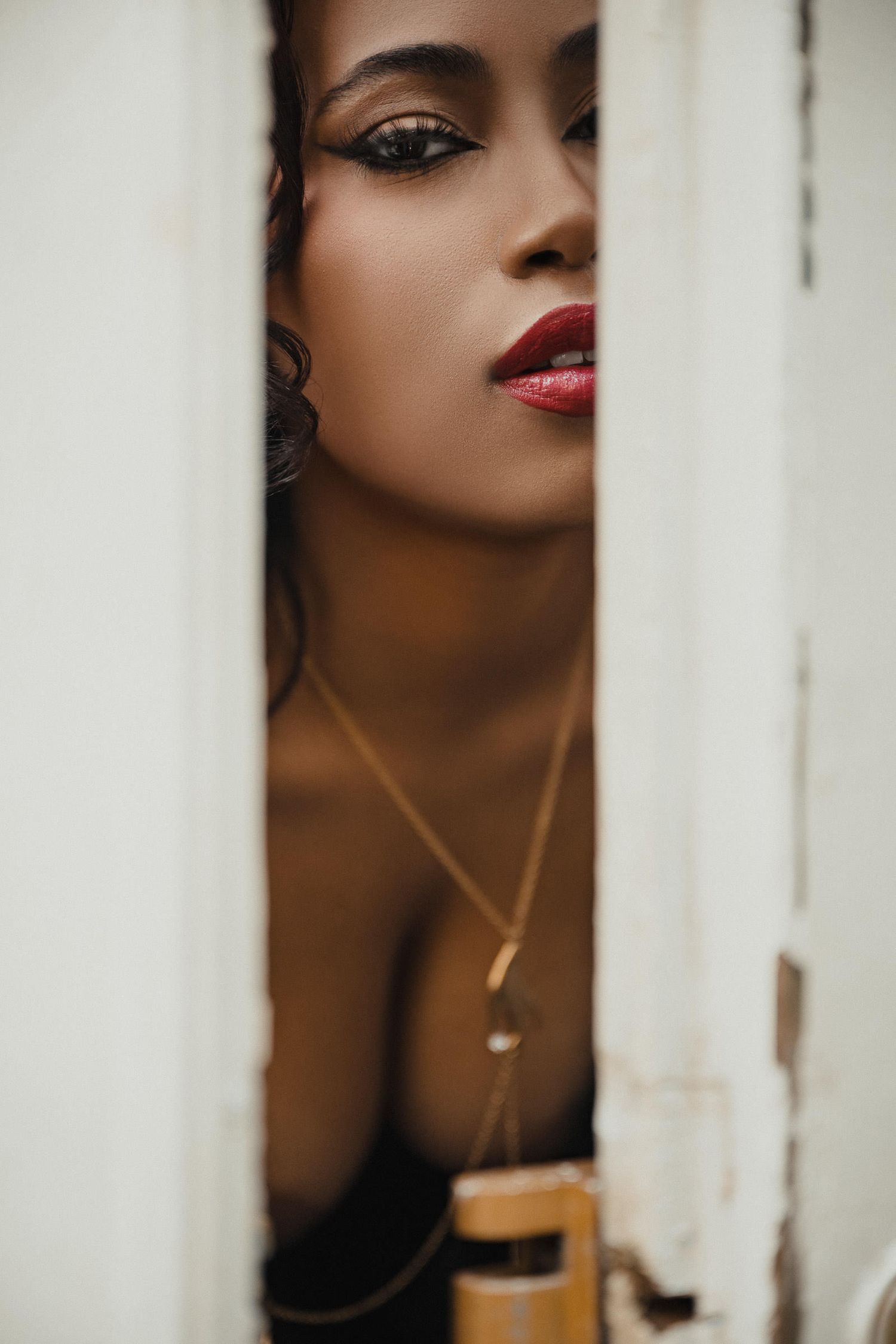 A close-up artistic portrait through white bars showing red lipstick and gold necklaces against dark skin. fun boudoir photoshoot ideas