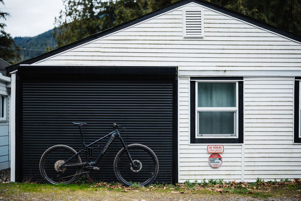 Black mountain bike leaning against white garage in small mountain town.