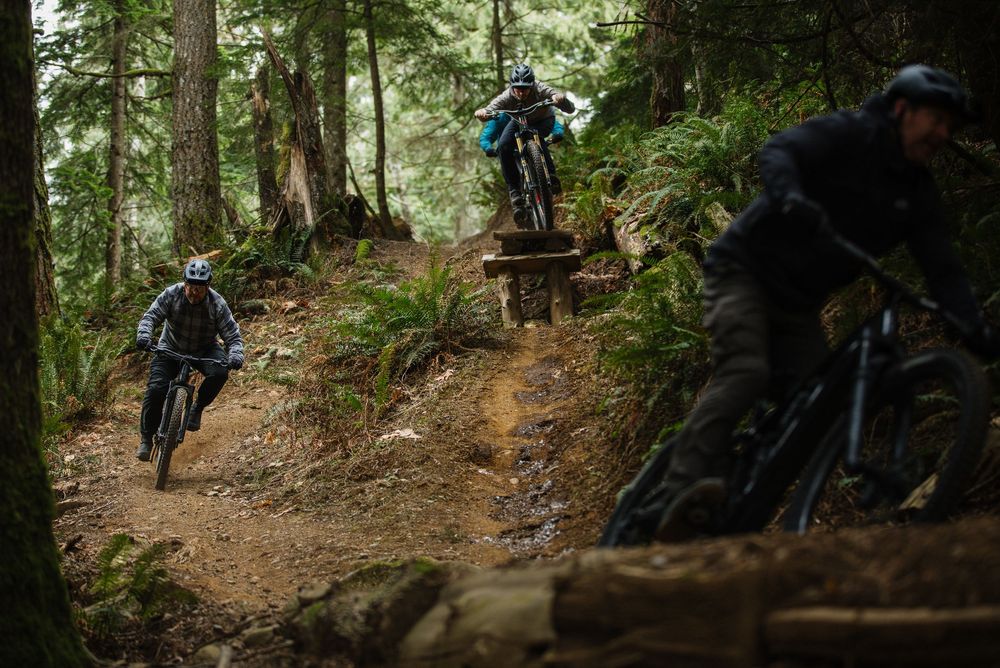 Mountain bikers navigating a rocky wooded trail with wooden features in dense forest.
