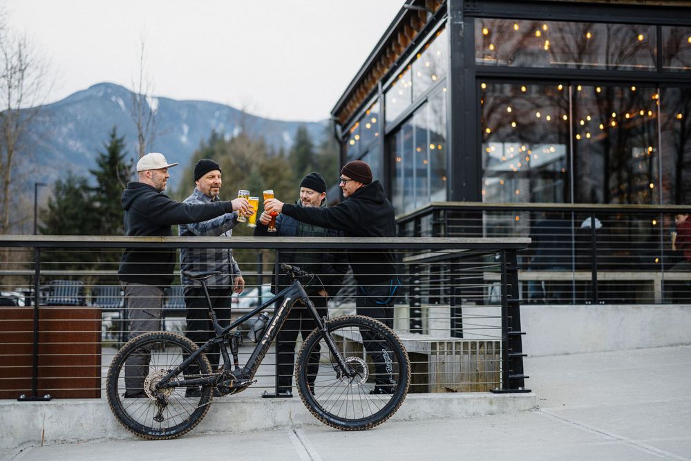 Group of friends toasting drinks beside a black mountain bike on an outdoor patio with mountain views.