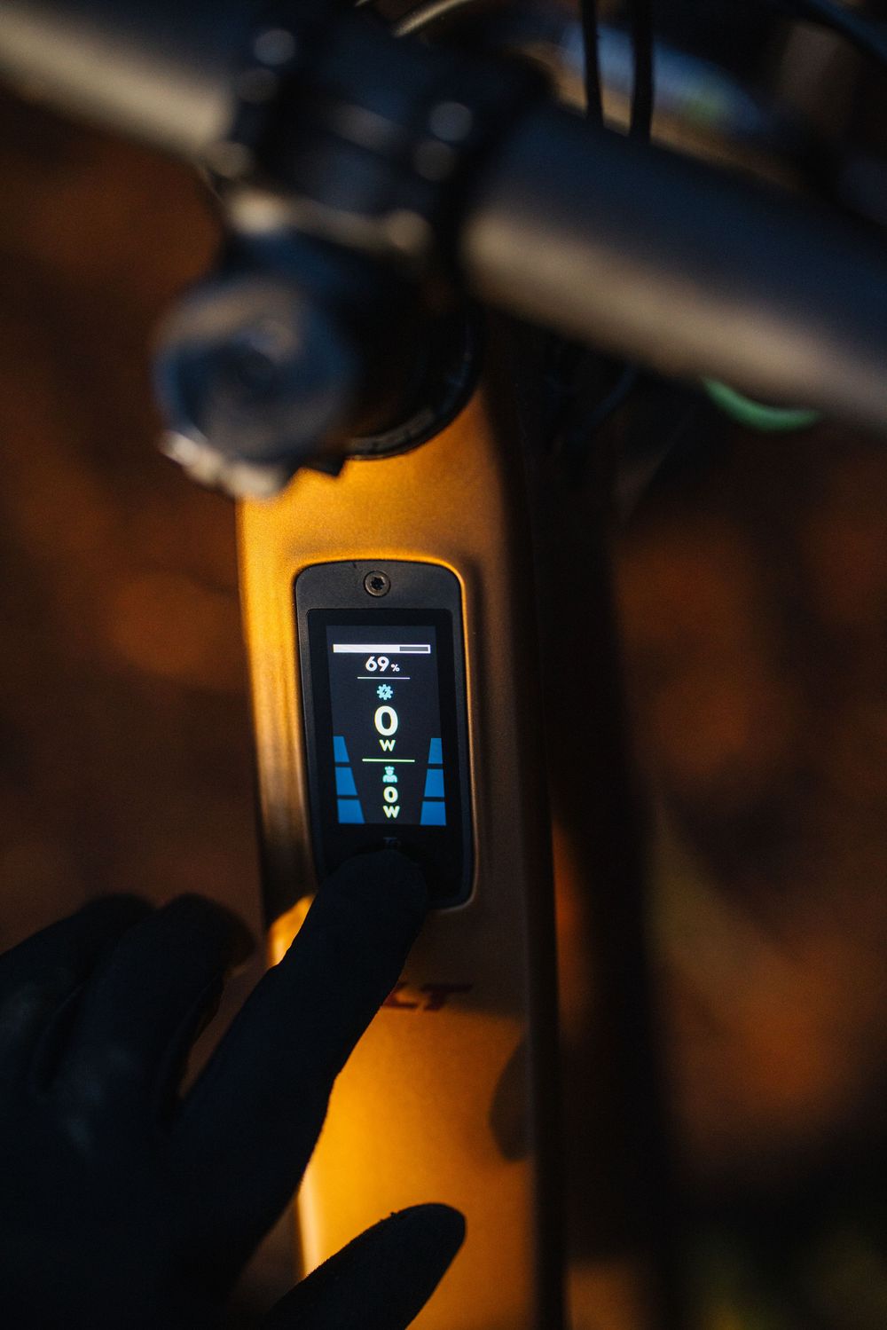 Gloved hand touching the display screen of an orange electric mountain bike showing battery and power metrics.
