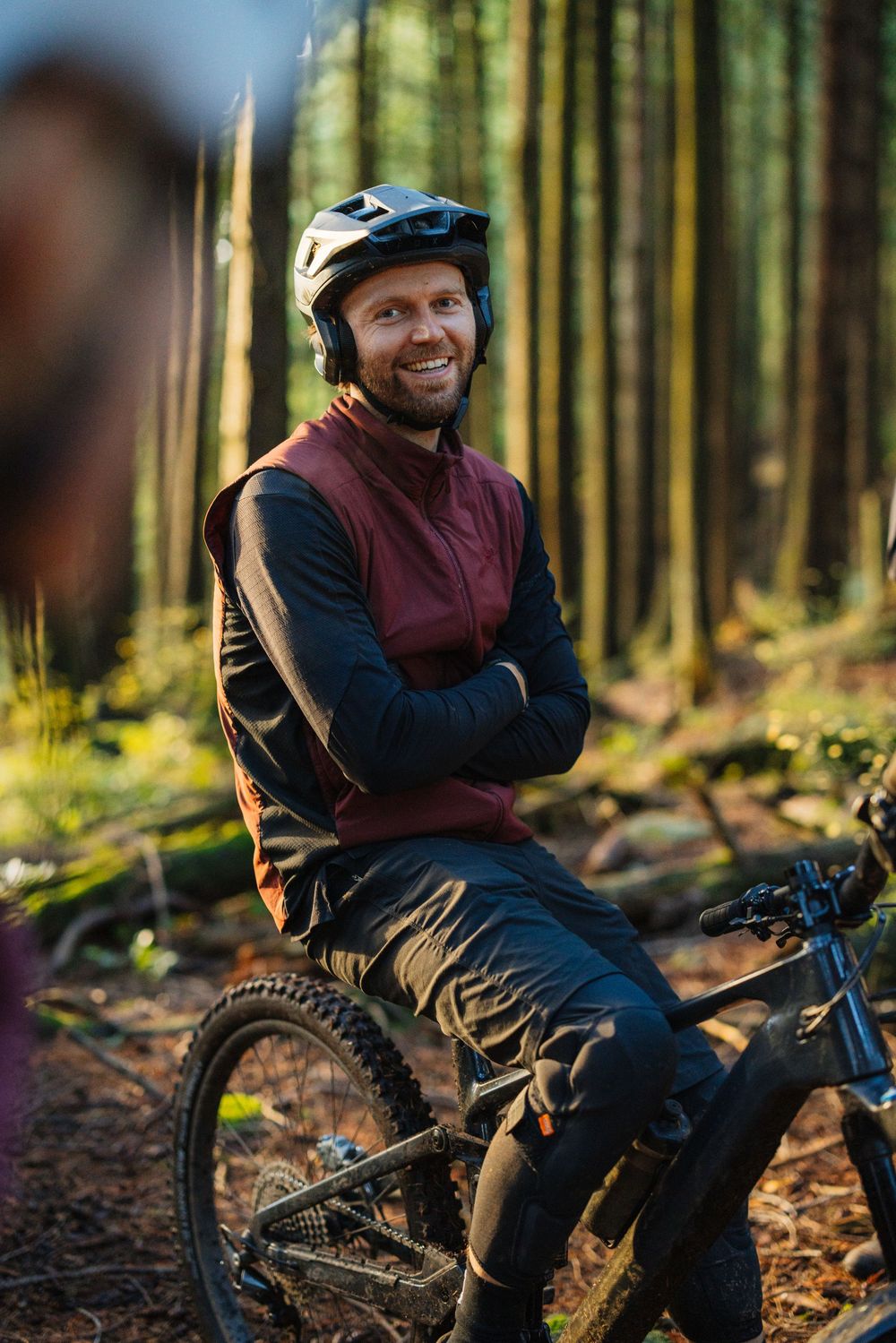 Smiling male mountain biker in helmet and gear resting on bike in sunlit forest trail.