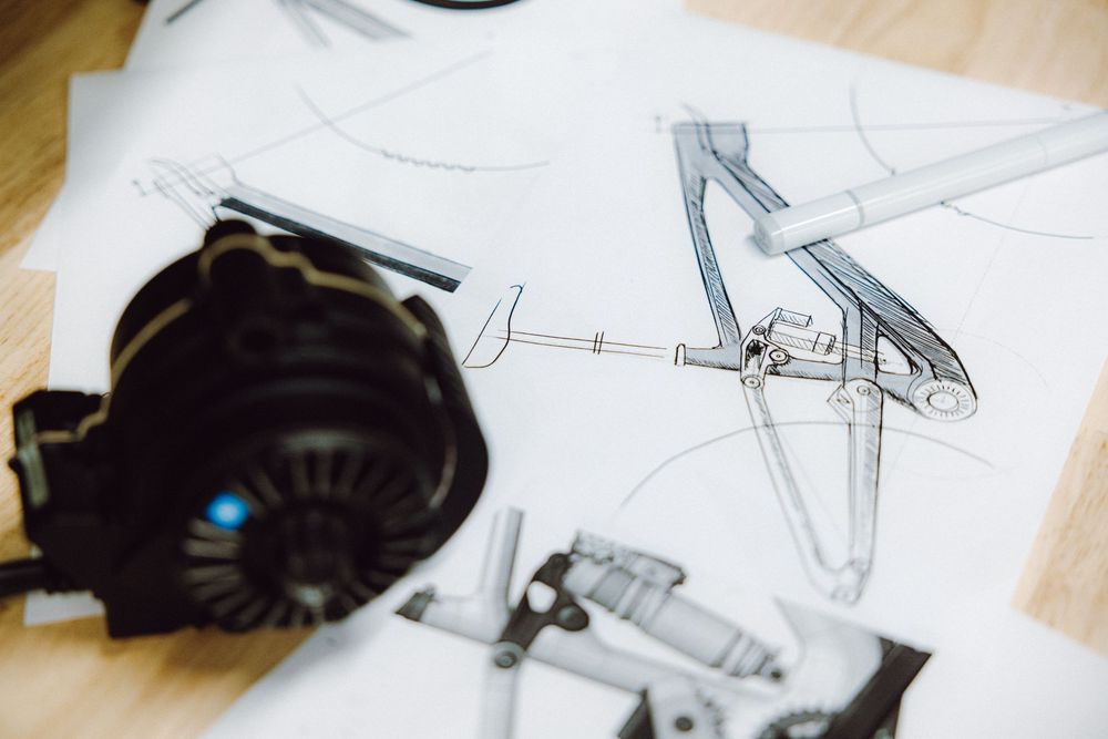 Technical pencil sketches of a bicycle frame design with drawing compass and camera lens on paper.