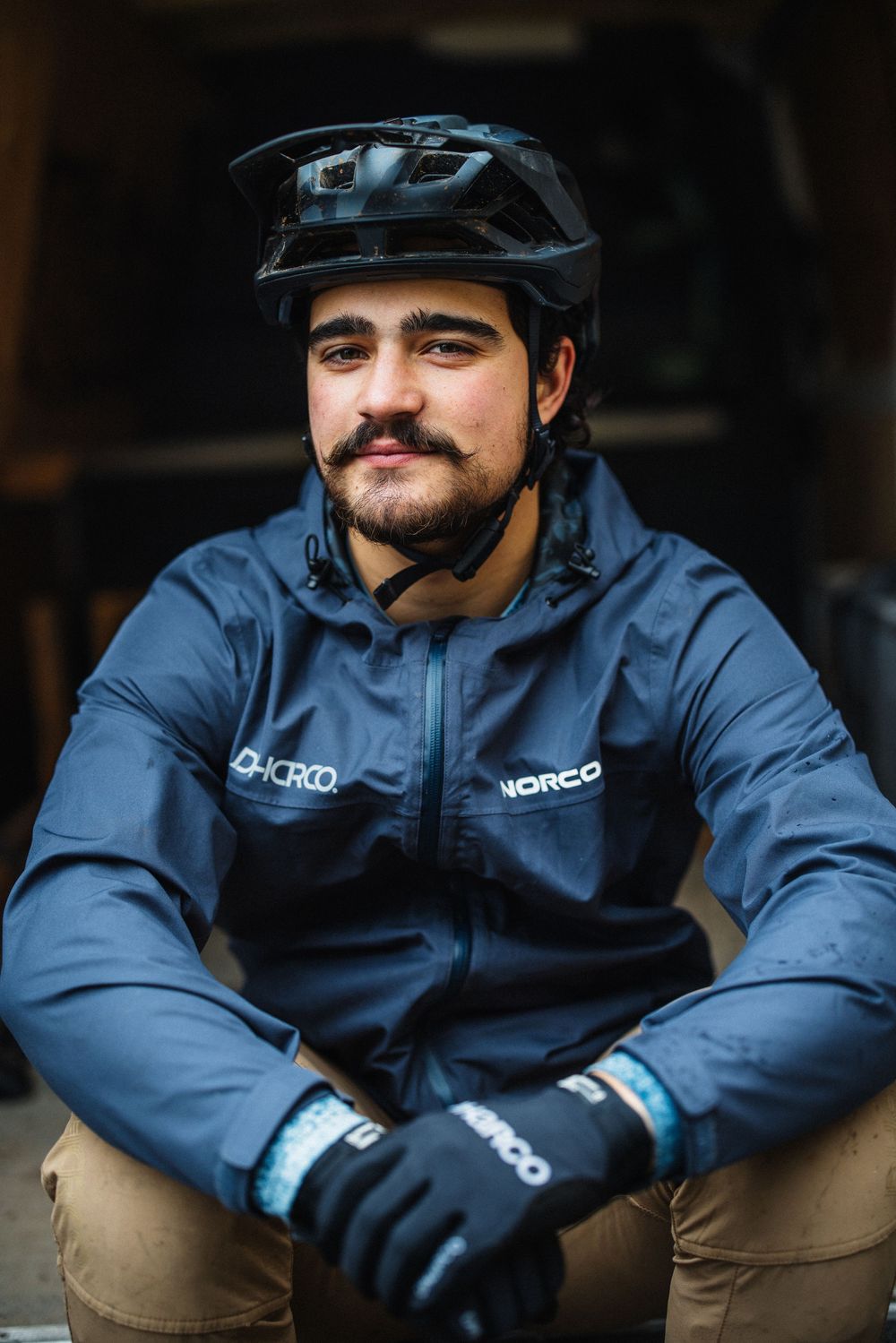 A male mountain biker wearing a Norco jacket and helmet sits relaxed in a garage setting.