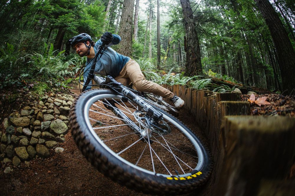 Mountain biker leaning sharply into a forest trail turn on a full-suspension bike among towering redwood trees.