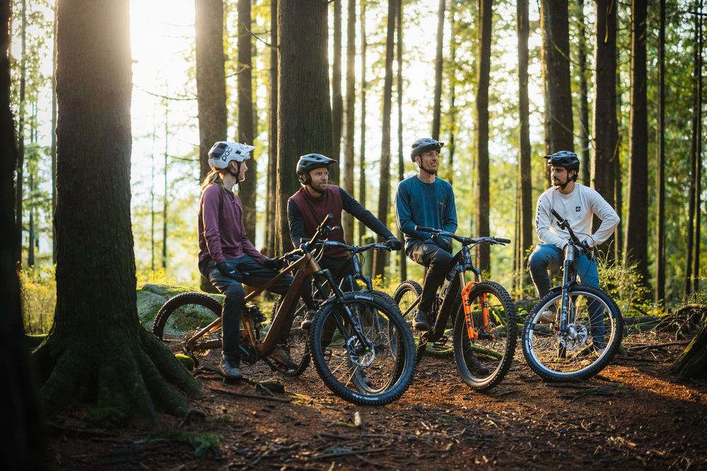Group of four mountain bikers with helmets gathered in sunlit forest trail.