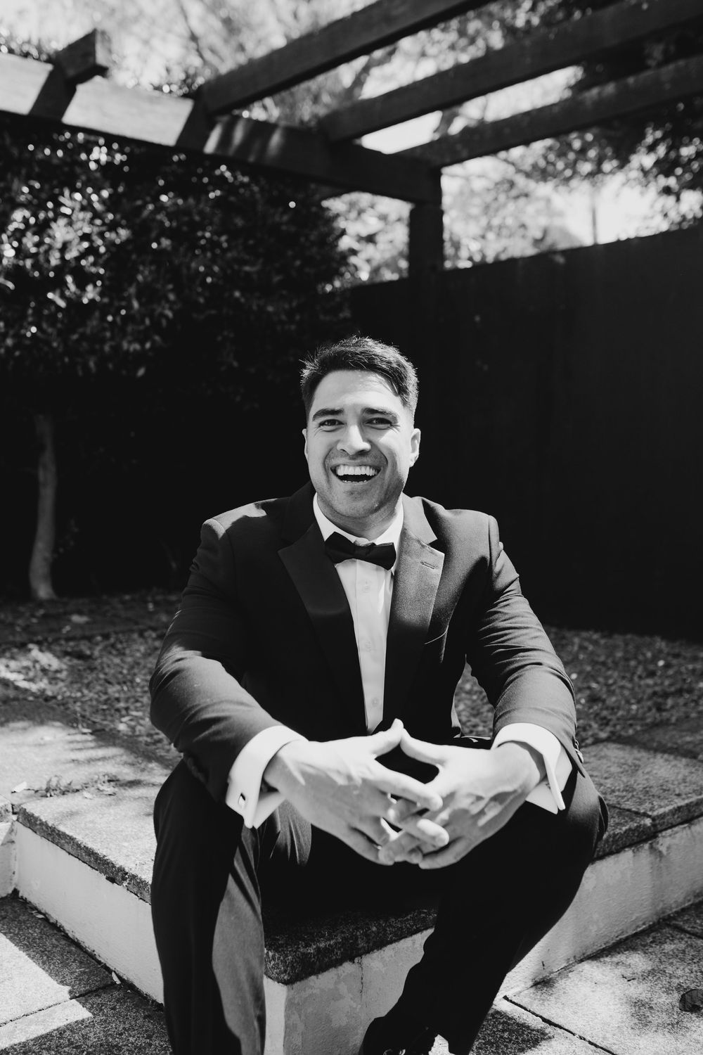 Smiling groom in tuxedo with bow tie sitting outdoors in black and white wedding portrait.