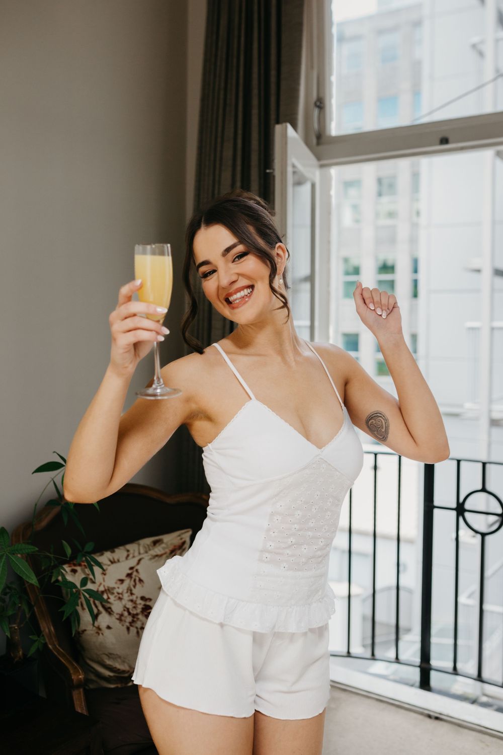 Happy bride in white pajamas holding champagne glass celebrating near window before wedding.