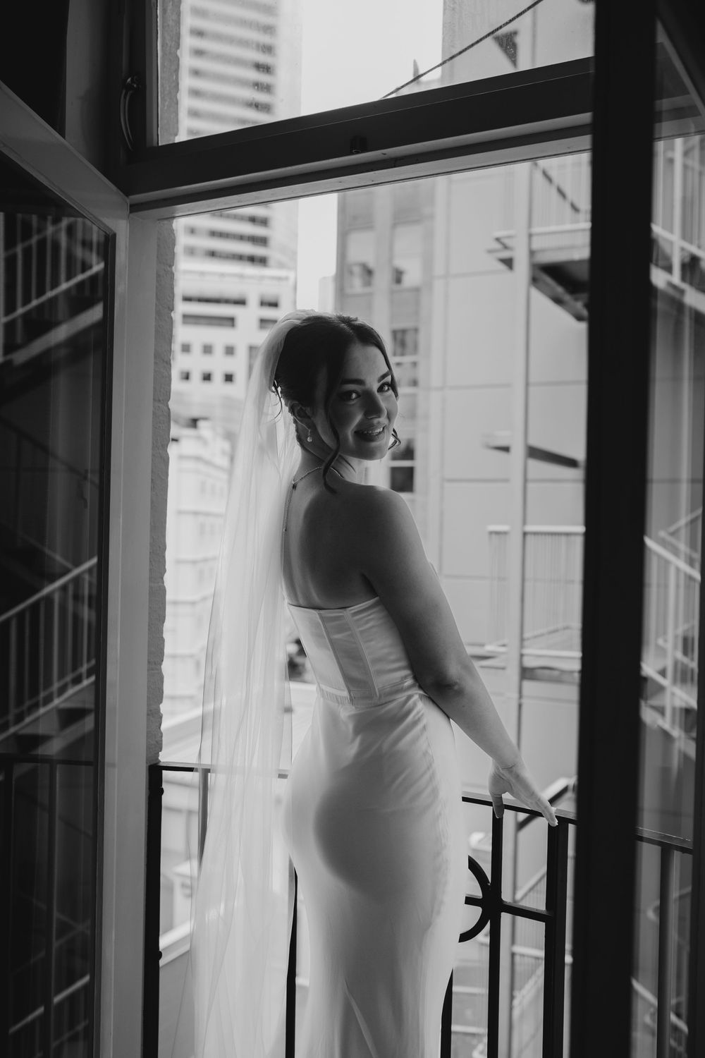 Bride in fitted white wedding gown and veil smiling on hotel balcony with city view.