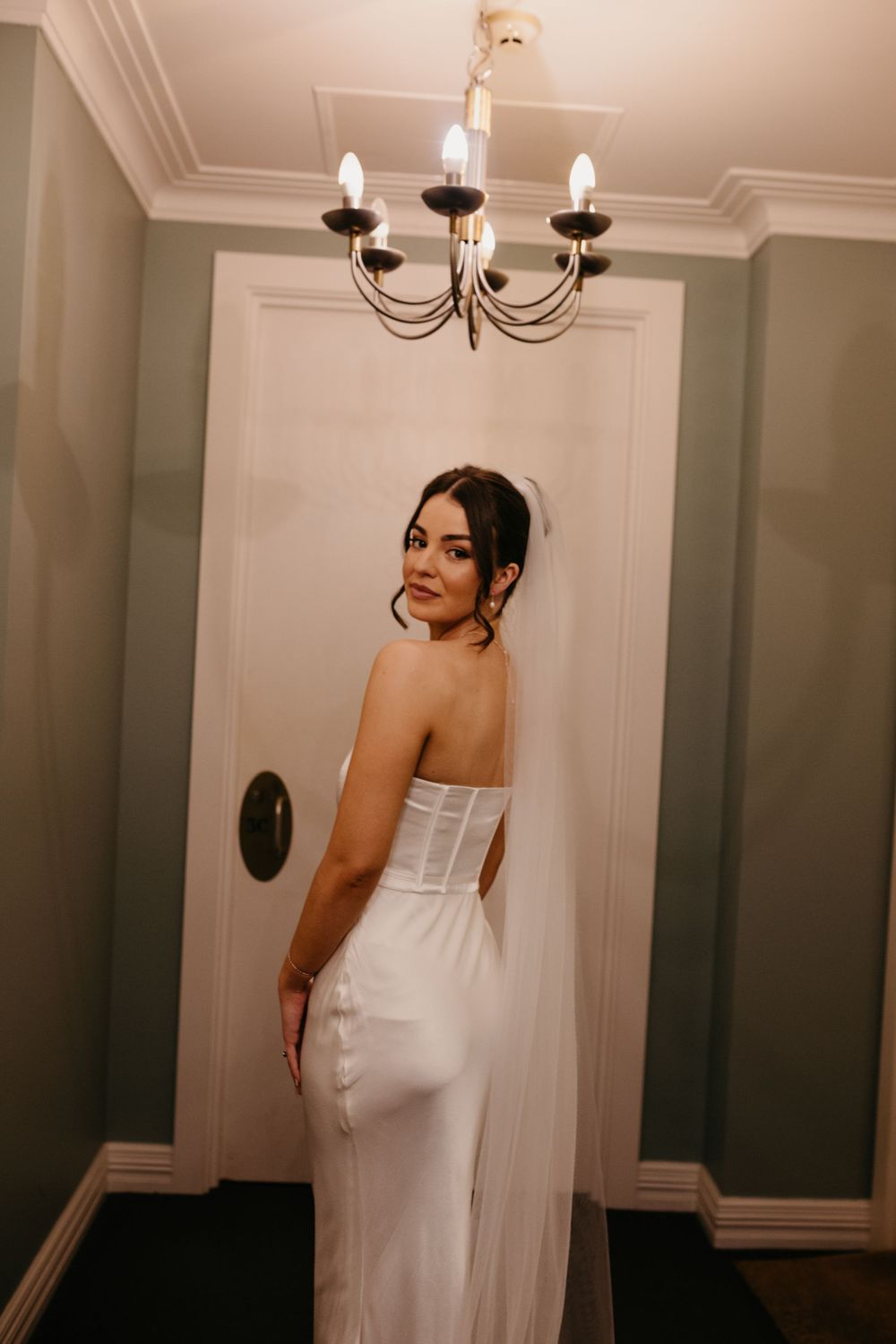 Bride in strapless white satin gown posing under chandelier in elegant green hallway.