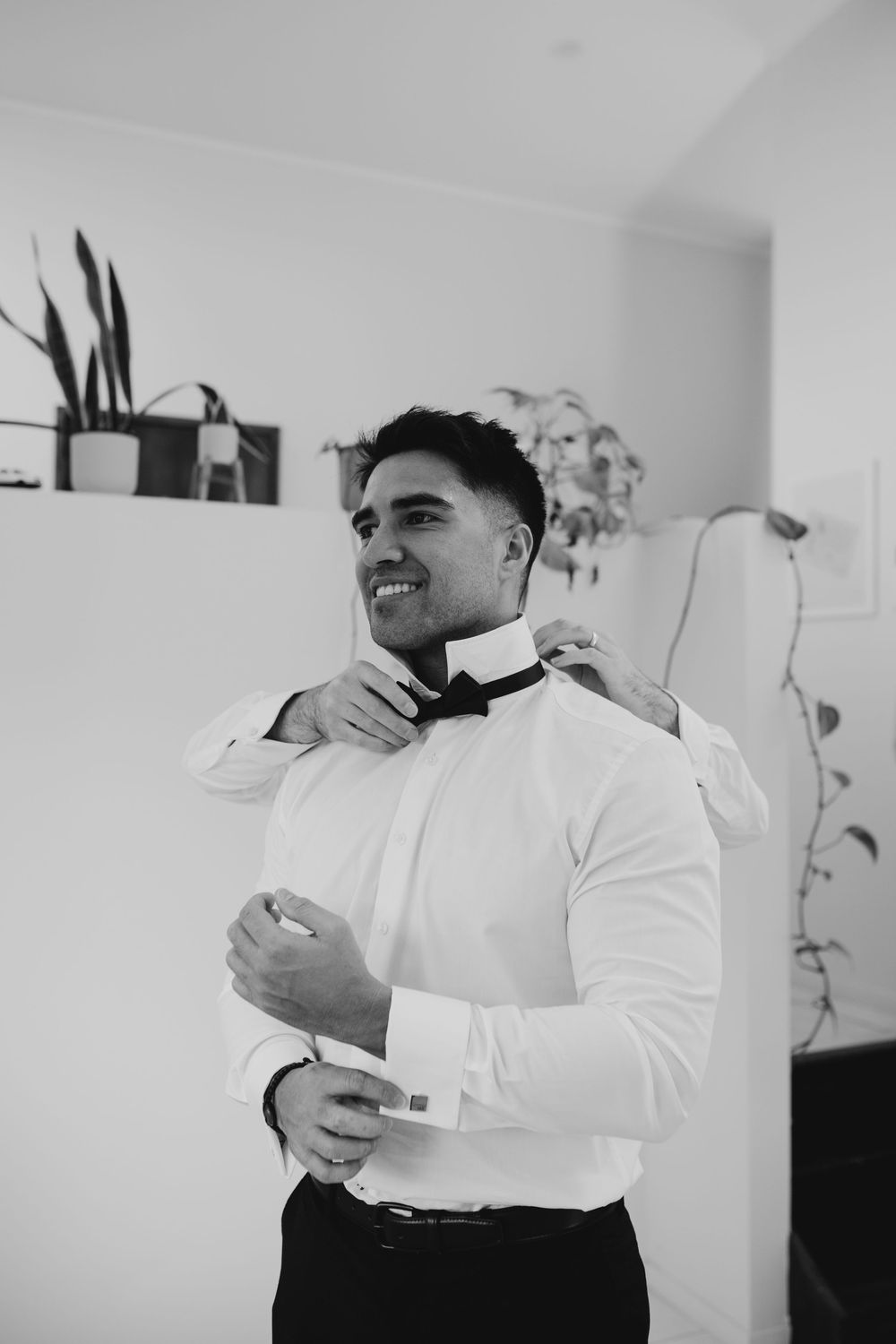 Black and white photo of a smiling groom adjusting his bow tie while getting ready for his wedding.
