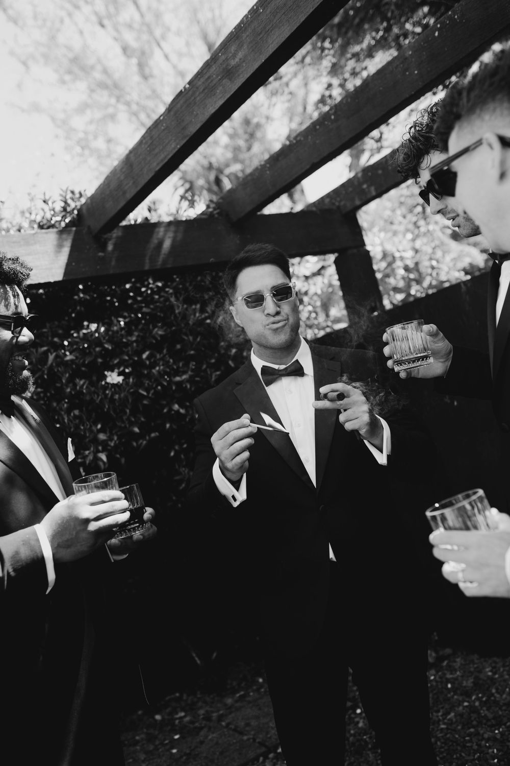 Black and white photo of a groom with groomsmen celebrating with drinks and cigars outdoors.