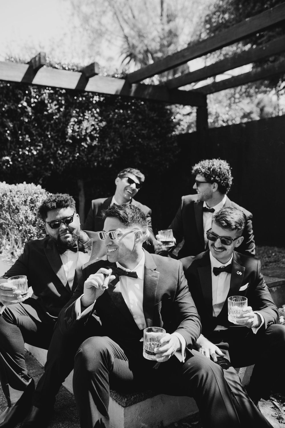 Five groomsmen in tuxedos celebrating with cigars and whiskey glasses outdoors in black and white photo.