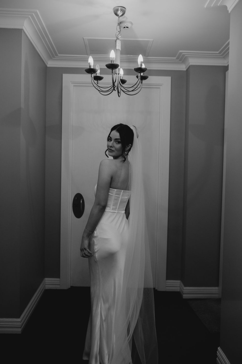 Elegant bride in strapless white gown with veil standing beneath chandelier in black and white photo.