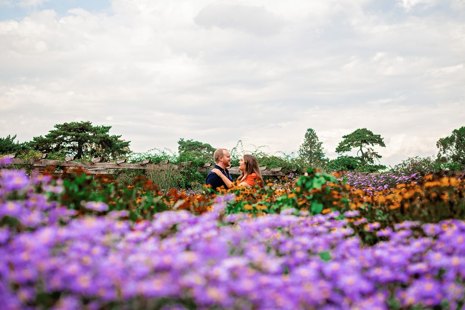 Couple sitting together in a vibrant purple flower field with lush green trees and cloudy sky backdrop.