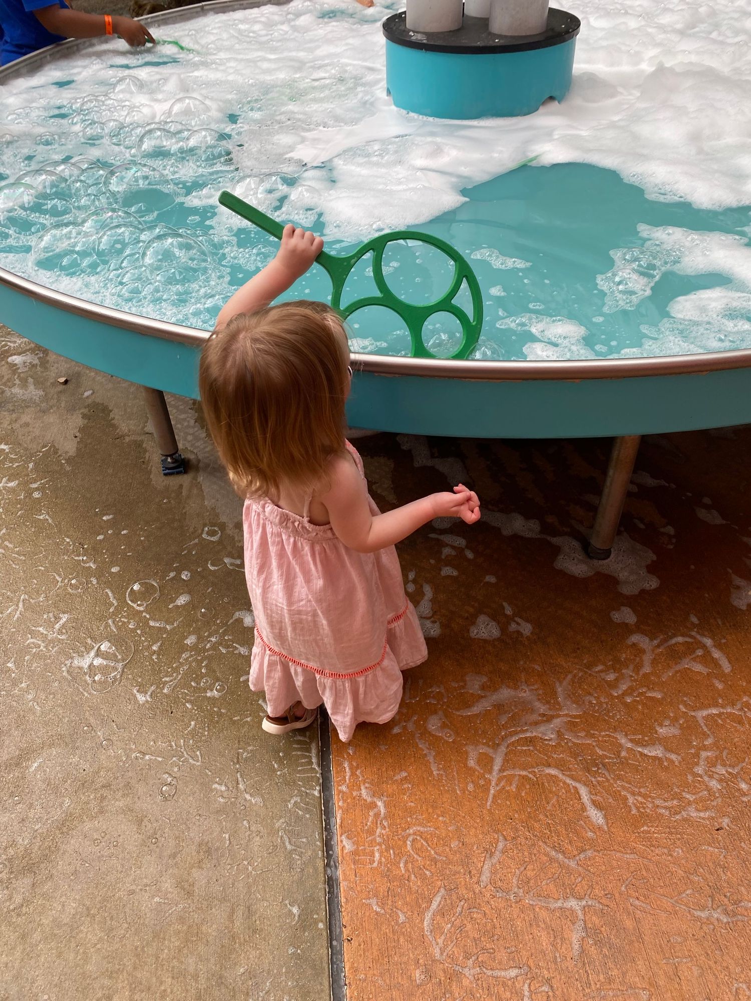 Toddler girl in pink dress playing with bubble foam at a water table activity station.