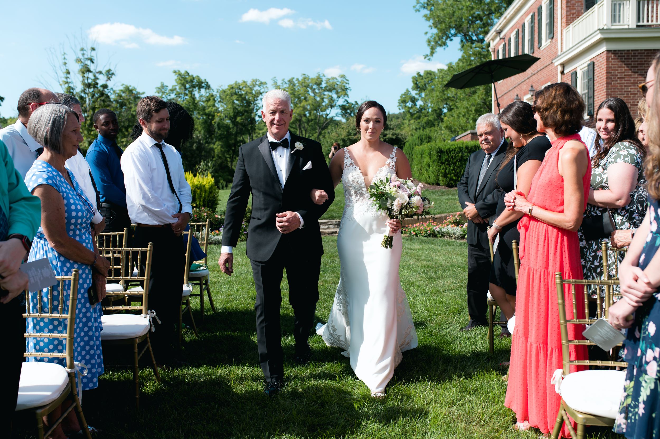 Bride in white gown walks down outdoor aisle with escort at sunny wedding ceremony.