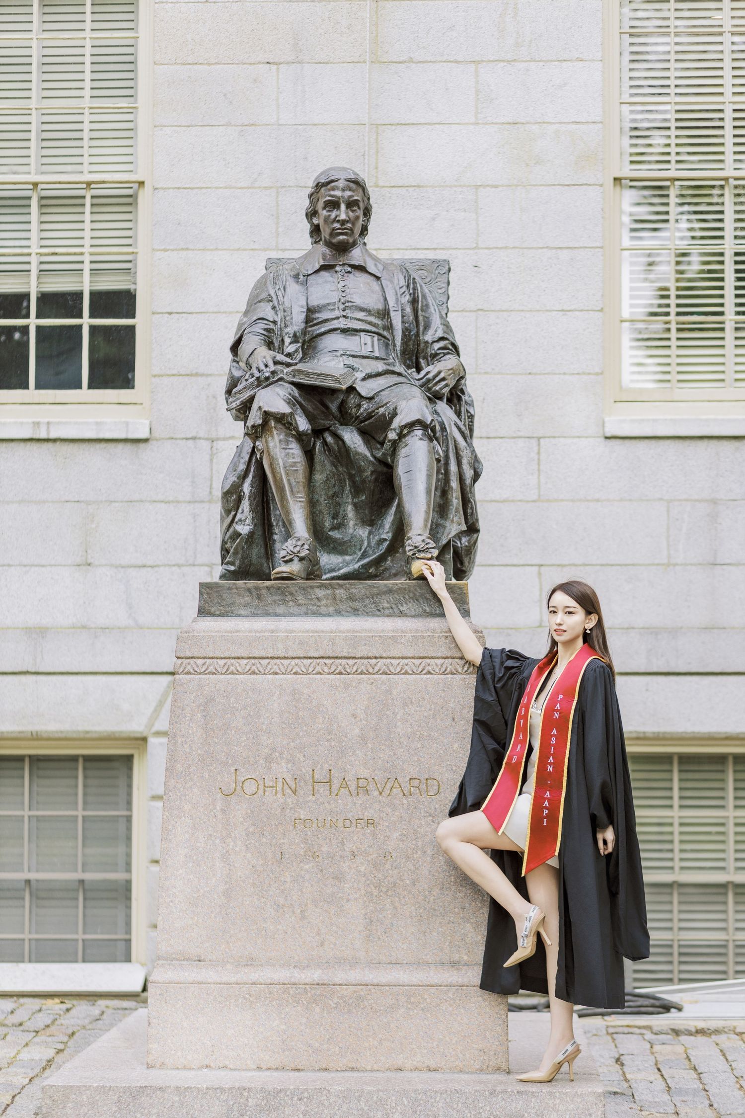 Graduate in cap and gown posing beside the John Harvard statue at Harvard University.