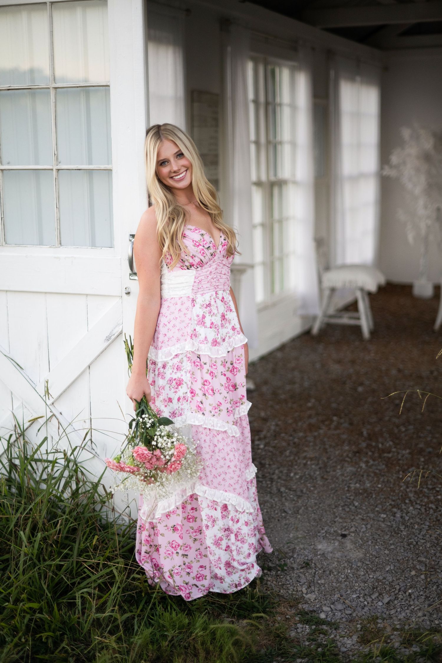 A smiling redhead woman in a floral maxi dress poses joyfully in a woodland setting with bare trees.