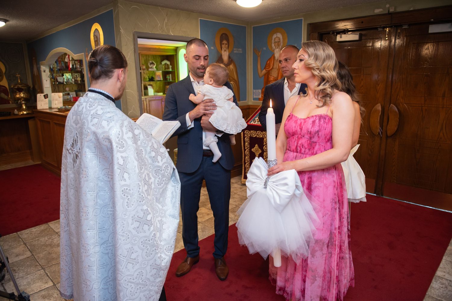 Orthodox priest greeting family with baby during christening ceremony inside decorated church.
