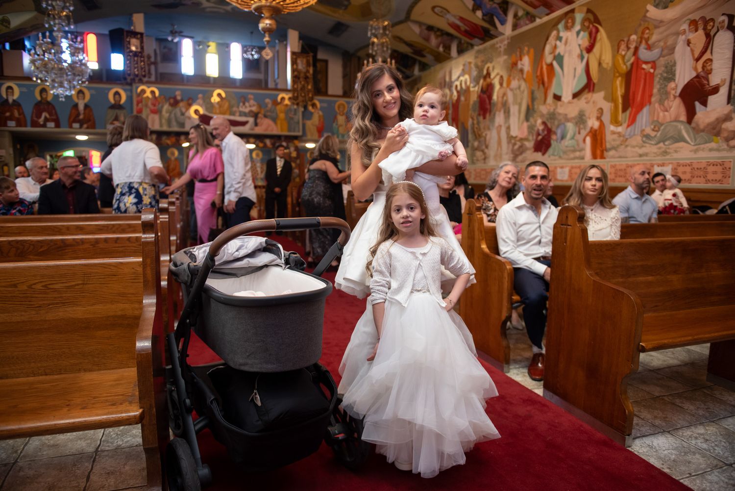 Mother holding baby in white baptism outfit with young girl in tulle dress inside Orthodox church.