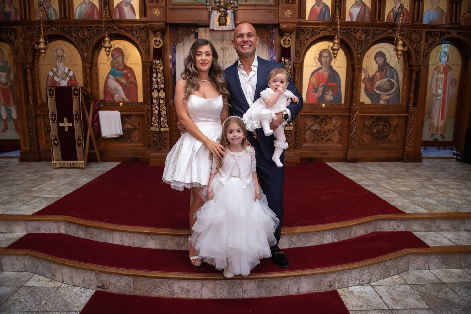 Family portrait at Orthodox church altar with parents, young girl in tulle dress, and baptism baby.