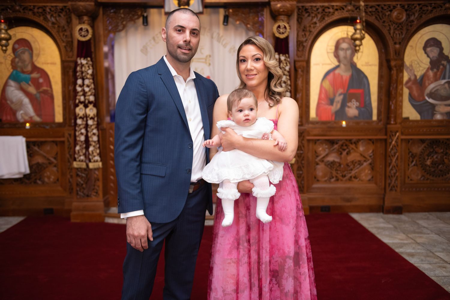 Couple in Orthodox church holding baby dressed in white baptism gown before iconostasis.