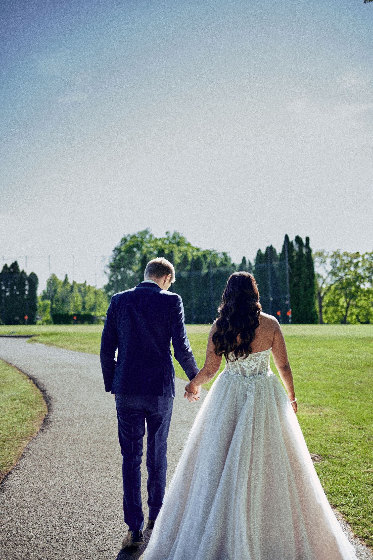 Bride and groom walking hand in hand down a path in a lush green park on their wedding day.