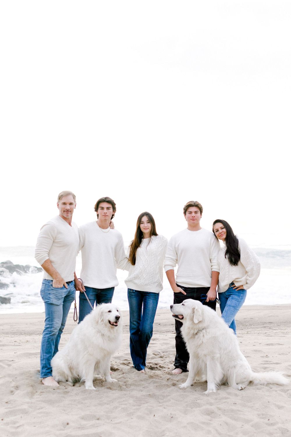 Family of five in white sweaters and jeans poses with two dogs on a sandy beach.