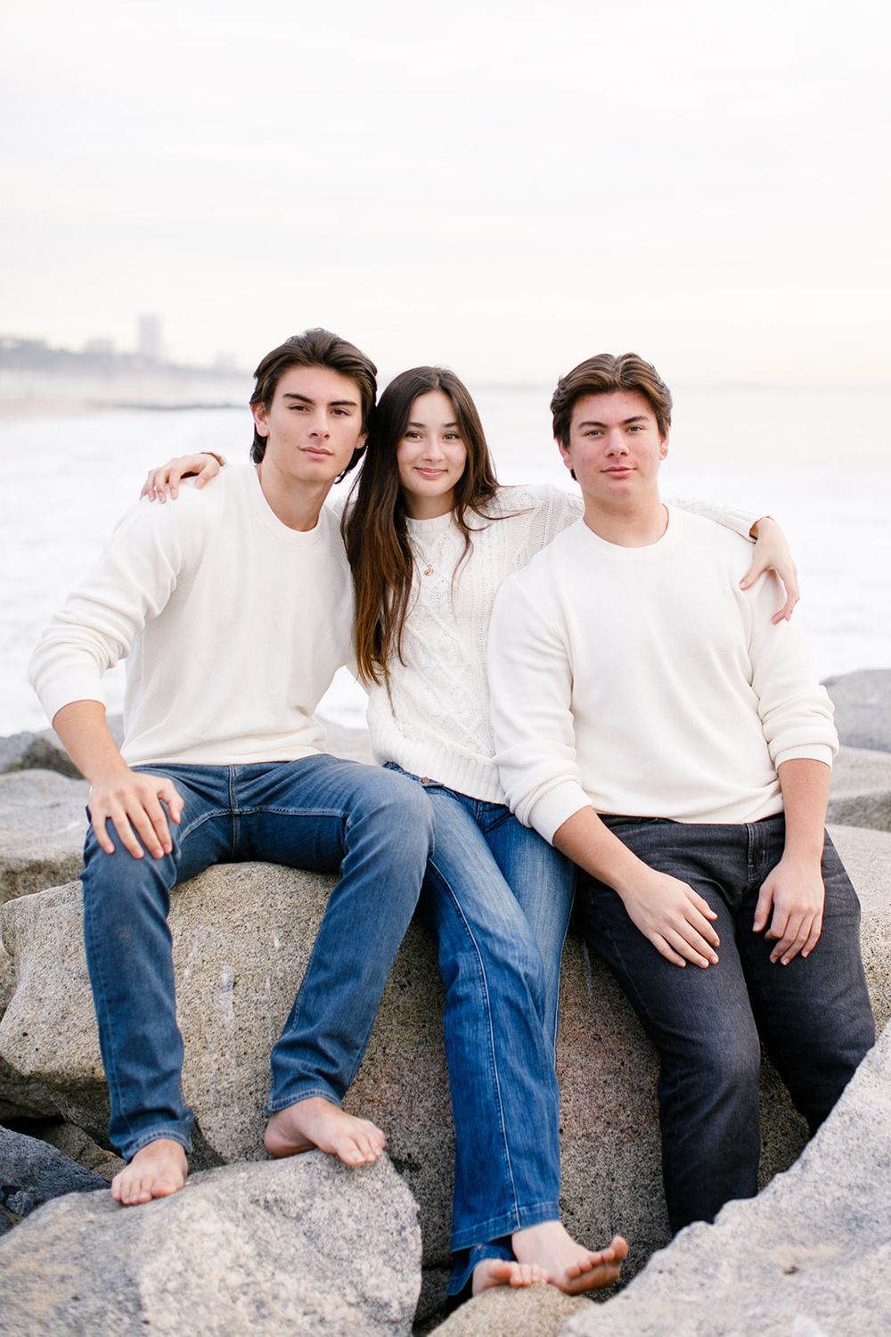 Three young adults in white sweaters and jeans sit on coastal rocks by the ocean, smiling warmly together.