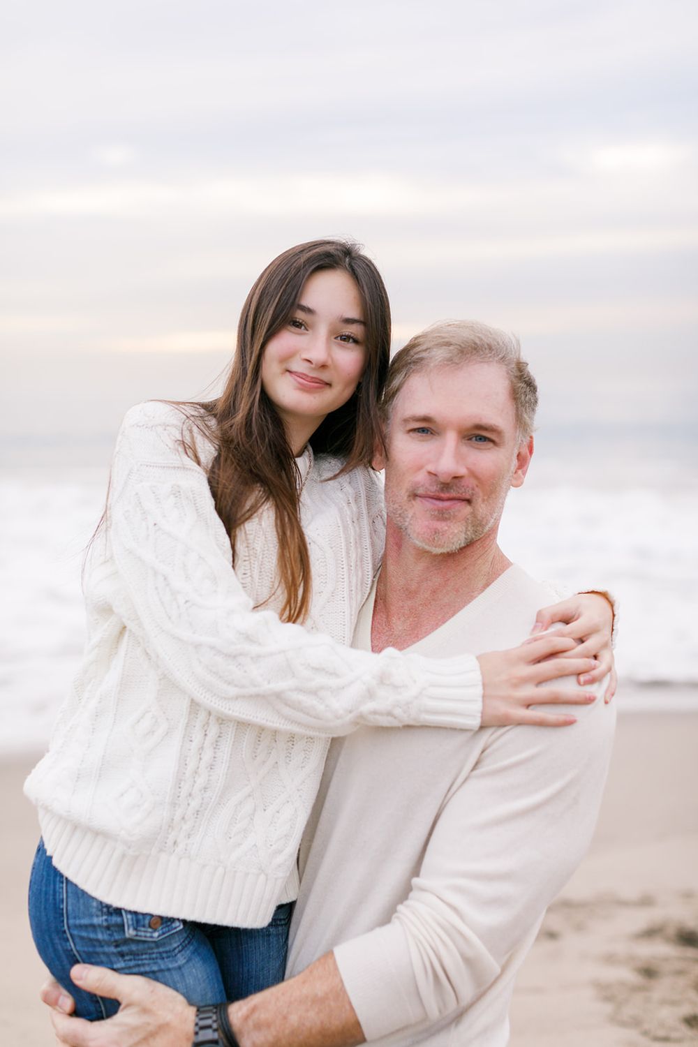 A couple embraces on a beach, both wearing white sweaters, with ocean waves in the background.