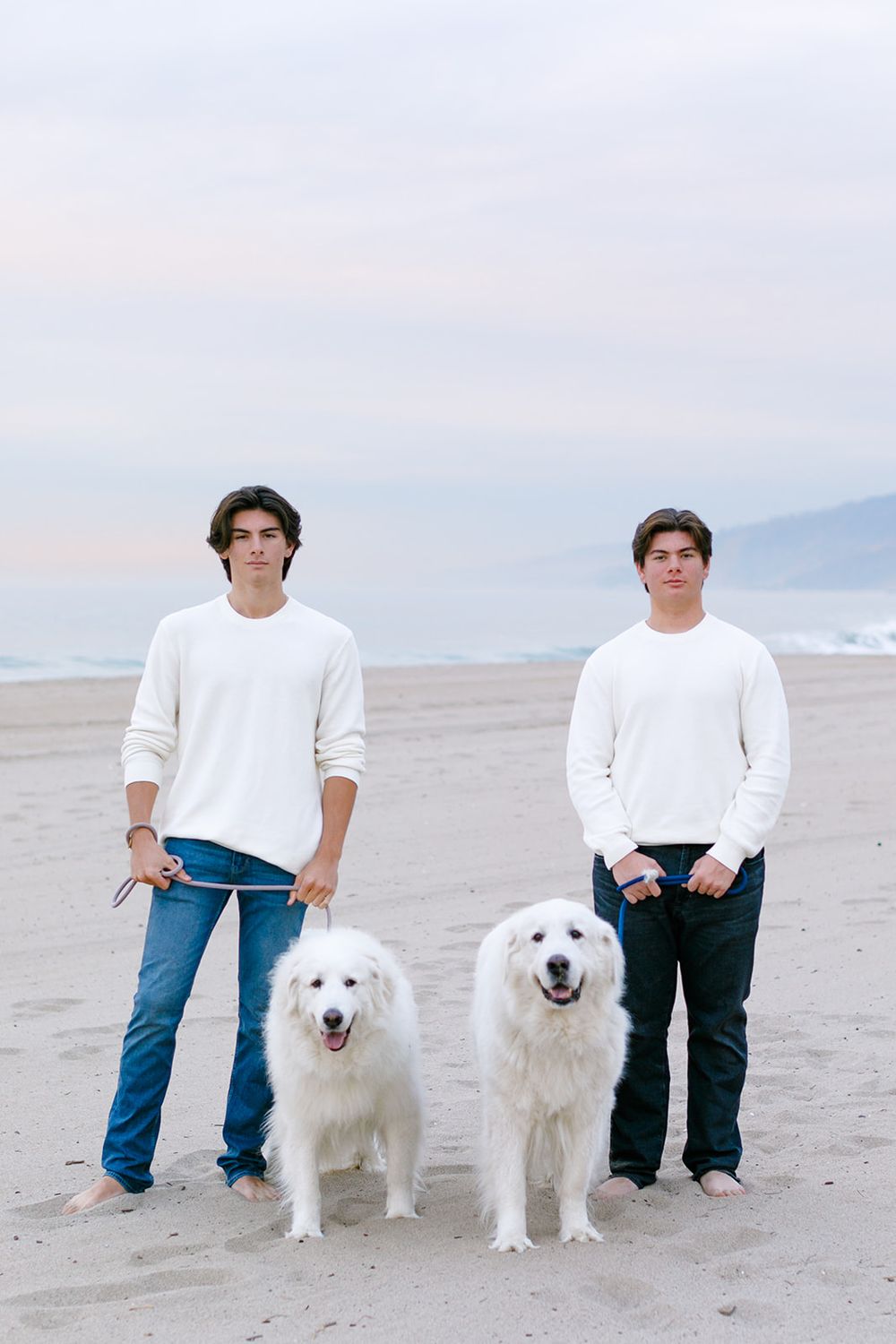 Two young men in white sweaters and jeans stand with two fluffy white dogs on a sandy beach at dusk.