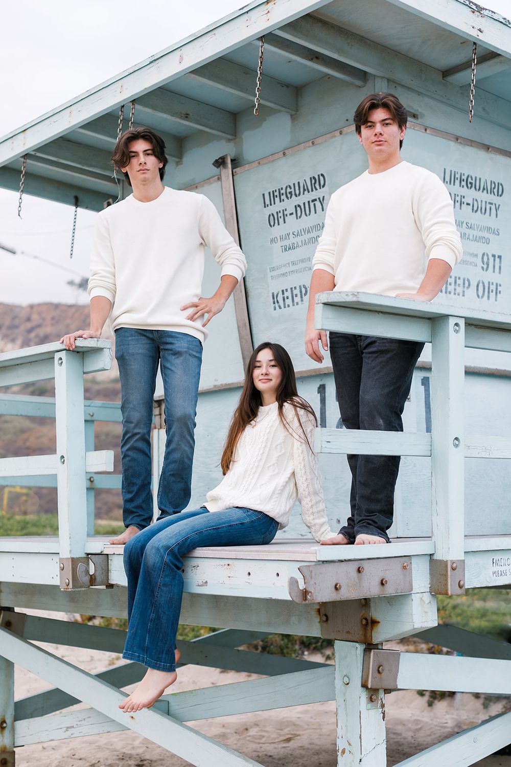 Three teens pose at a lifeguard off-duty station on the beach, two boys standing and a girl sitting.