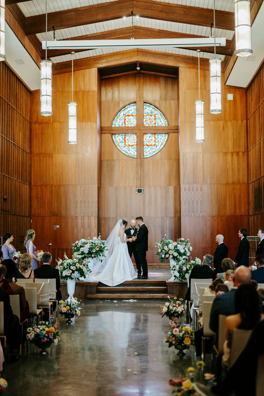 Bride and groom exchange vows at chapel altar with stained glass cross, floral arrangements, and wood-paneled walls.